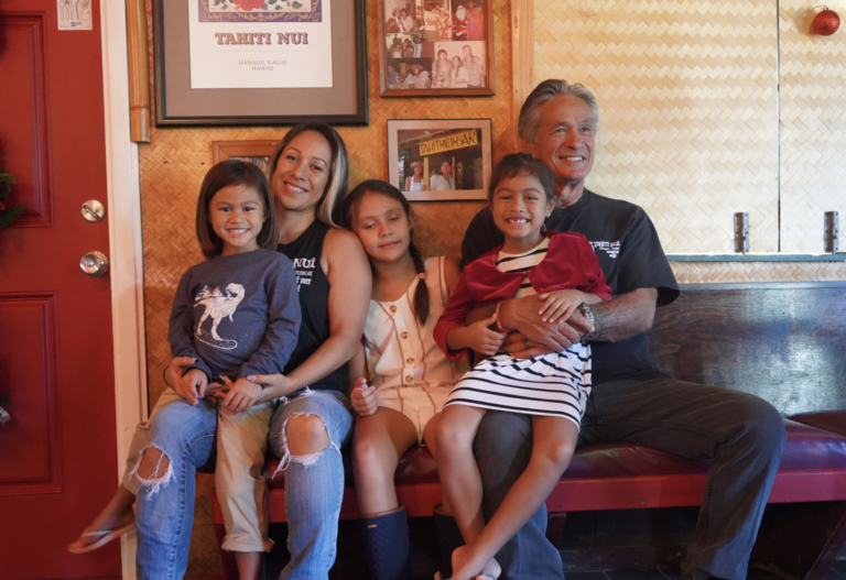 A family of 5 sit on a bench in a room decorated with framed Tahiti Nui Memorabilia.