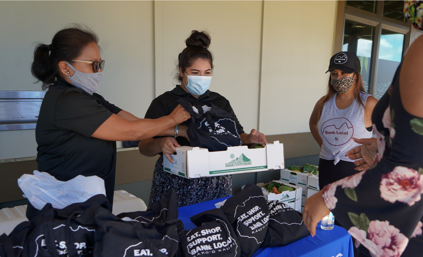 Two women filling box with fresh produce to give to a community member. Filled canvas totes on the table with a message to eat, shop, support, bank local.