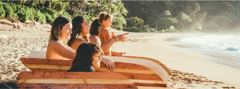 two women and three children sit on the beach with their beautiful wooden surfboards. One woman points out to the water and everyone looks out to see what she sees.