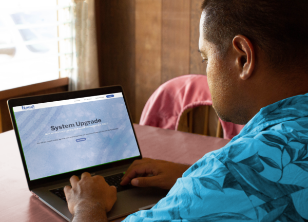 A man in a teal Hawaiian shirt sits with a laptop. The screen shows the Kauai FCU website on a page explaining the system upgrade.